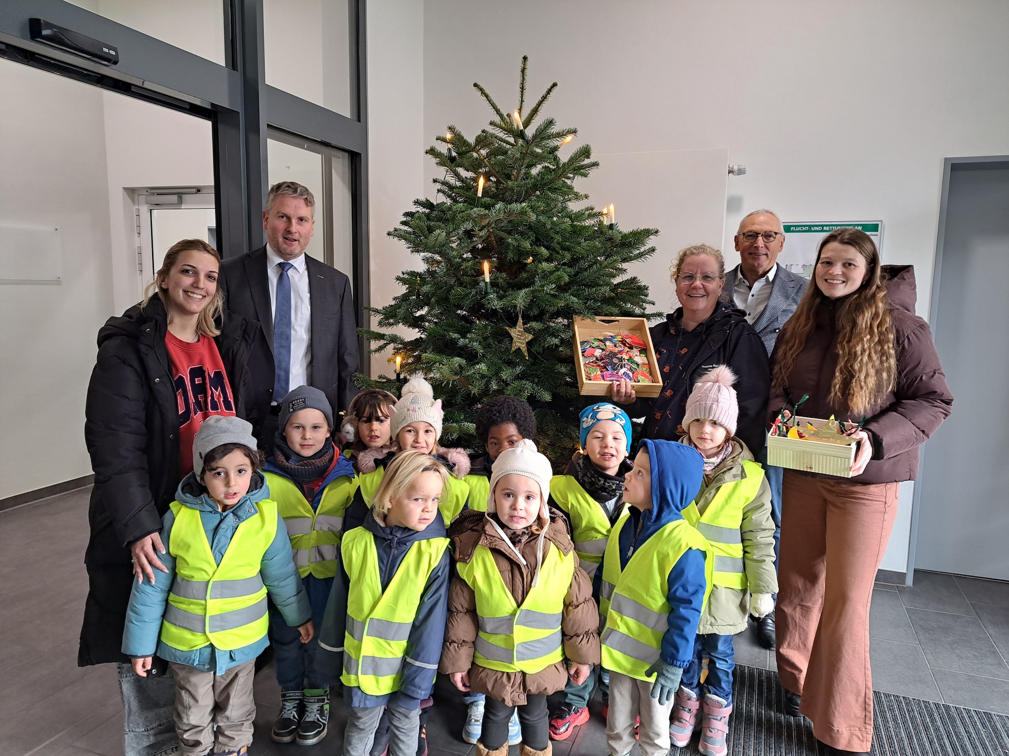 Kinder in gelben Warnwesten, drei Frauen sowie zwei geschäftlich gekleidete Männer stehen vor einem Weihnachtsbaum. 