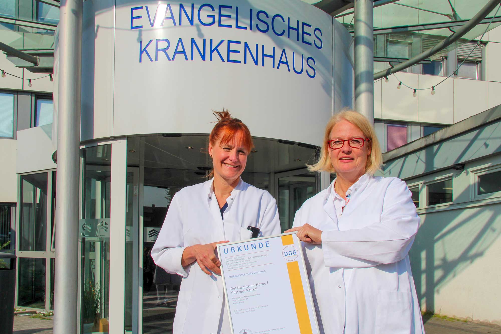 Zwei Frauen in weißen Kitteln halten eine Urkunde vor dem Evangelischen Krankenhaus, einem modernen Krankenhausgebäude.