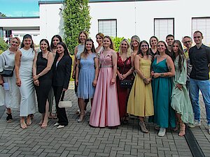 Eine festlich gekleidete Gruppe von jungen Menschen, die im Freien auf einem gepflasterten Platz stehen und in die Kamera lächeln.