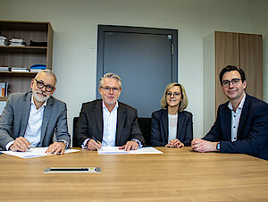 Drei Männer und eine Frau in Businesskleidung sitzen an einem Tisch. Zwei Männer von ihnen halten einen Stift in der Hand. Vor ihnen liegen Zettel auf dem Tisch.