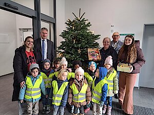 Kinder in gelben Warnwesten, drei Frauen sowie zwei geschäftlich gekleidete Männer stehen vor einem Weihnachtsbaum. 