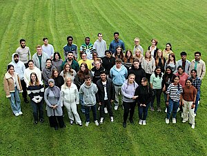 Eine Gruppe junger Menschen, die gemeinsam auf einer grünen Wiese stehen und für ein Foto posieren, von oben gesehen.