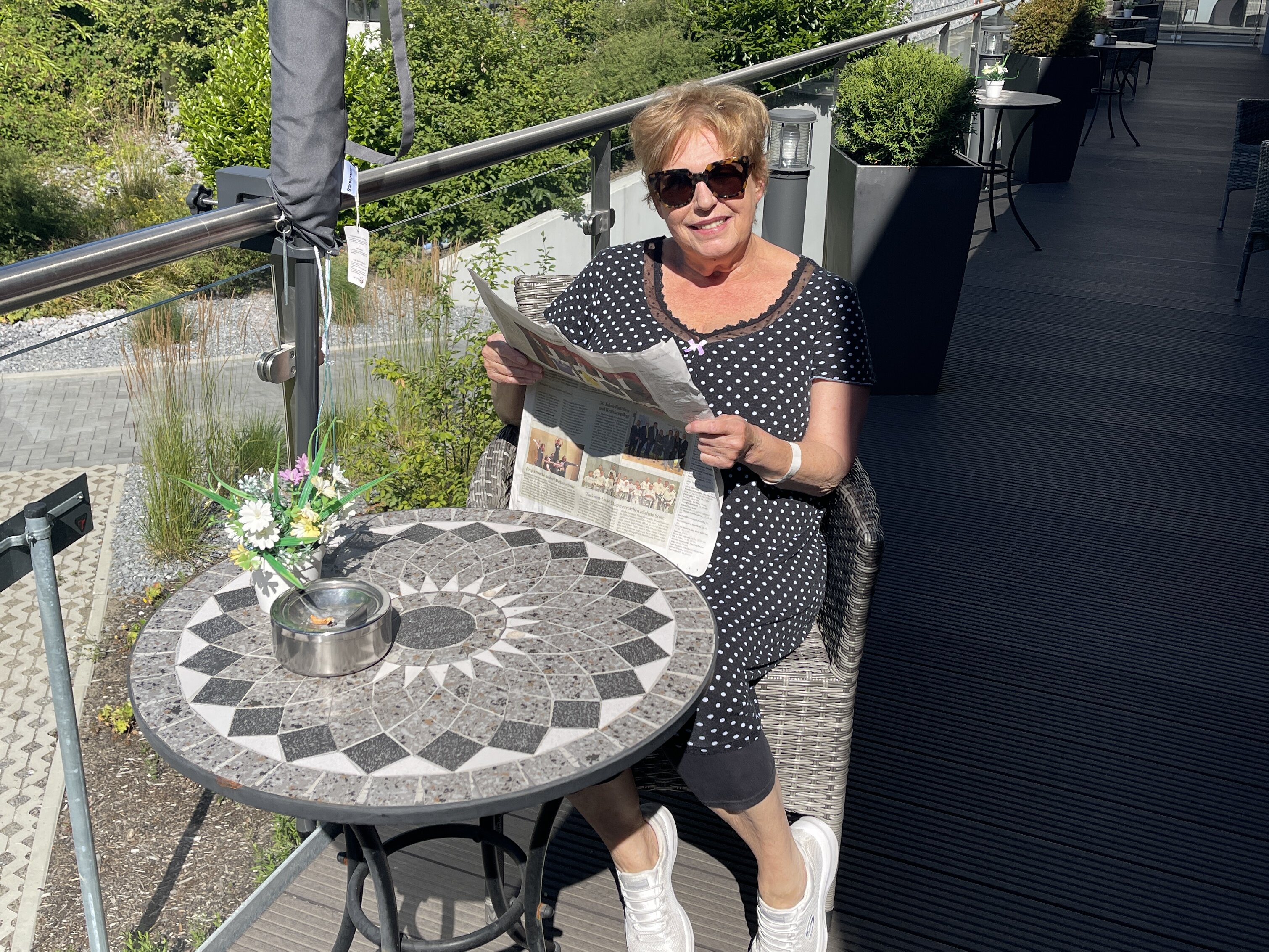 Eine Frau mit Sonnengrippe sitzt auf einem sonnigen Balkon und hält eine Zeitung in der Hand. 