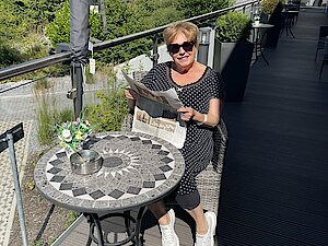 Eine Frau mit Sonnengrippe sitzt auf einem sonnigen Balkon und hält eine Zeitung in der Hand. 