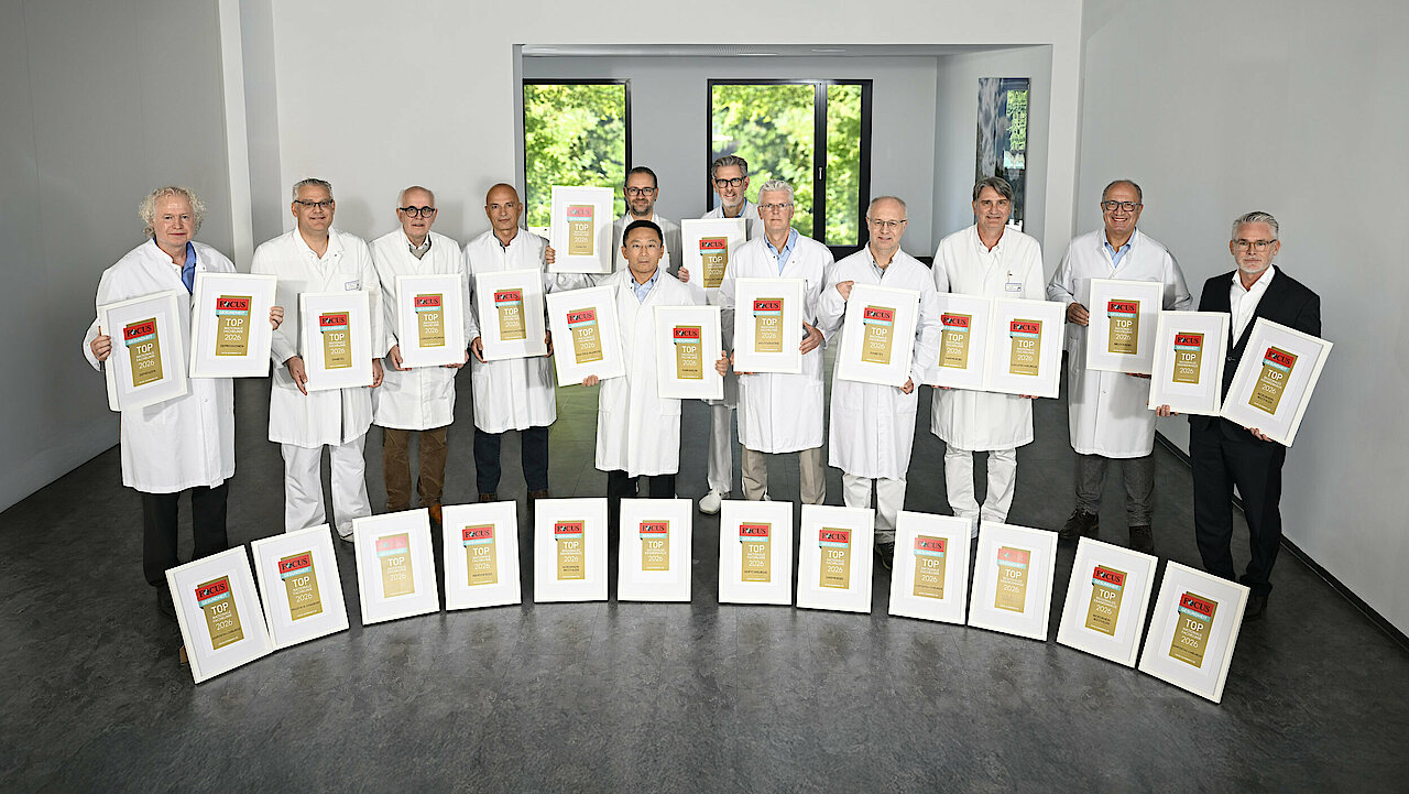 Viele Männer stehen nebeneinander. Bis auf ein Mann tragen alle weiße Kittel. Rechts außen steht ein Mann mit Brille und Bart im dunklen Anzug. Alle halten gerahmte Siegel in den Händen. Weitere Rahmen stehen davor.  den in 