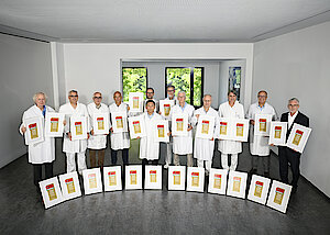 Viele Männer stehen nebeneinander. Bis auf ein Mann tragen alle weiße Kittel. Rechts außen steht ein Mann mit Brille und Bart im dunklen Anzug. Alle halten gerahmte Siegel in den Händen. Weitere Rahmen stehen davor. den in