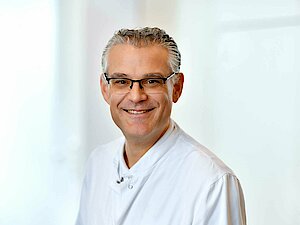 Ein lächelnder Mann mit grauem Haar und Brille, der einen weißen Arztkittel trägt, steht vor einem hellen Fenster.