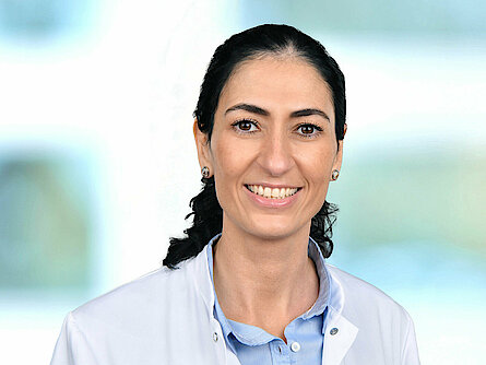 Frau mit dunklem Haar in einem Pferdeschwanz, weißem Laborkittel und blauem Hemd, lächelt in die Kamera.
