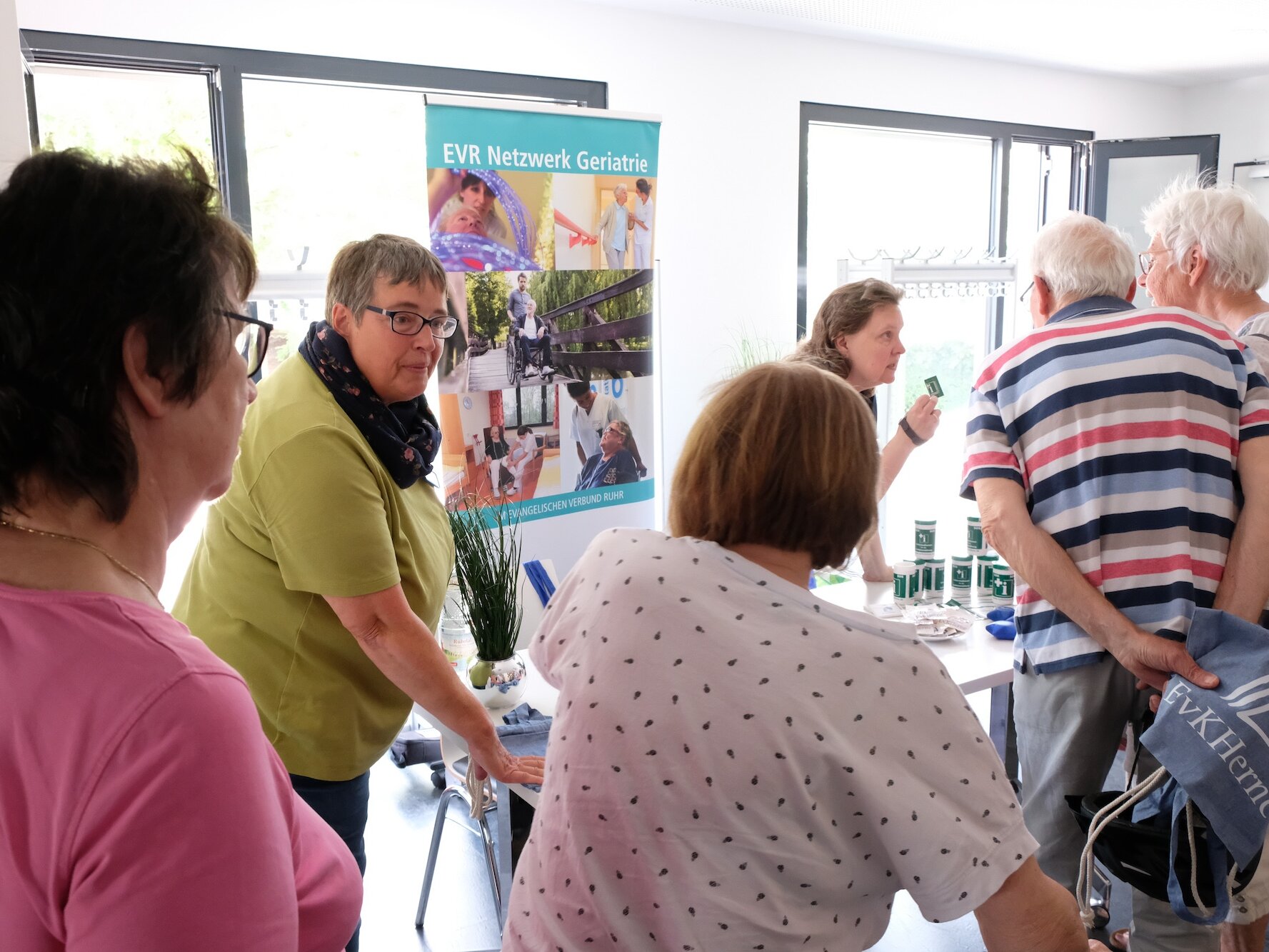 Mehrere ältere Menschen stehen in einem Raum, informieren sich an einem Stand. Eine Frau schiebt einen Rollator.