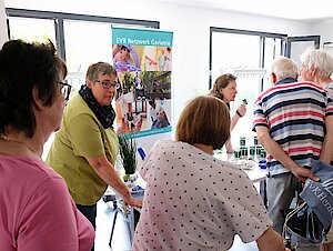 Mehrere ältere Menschen stehen in einem Raum, informieren sich an einem Stand. Eine Frau schiebt einen Rollator.