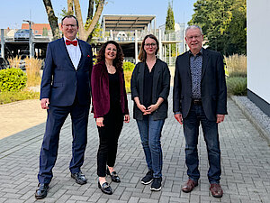 Vier Erwachsene stehen im Freien auf einer gepflasterten Fläche und lächeln in die Kamera, gekleidet in Geschäfts- und Freizeitkleidung.