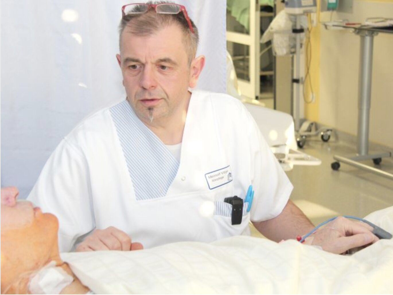 Ein Krankenpfleger im weißen Kittel kümmert sich aufmerksam um einen Patienten, der in einem Krankenhausbett in einem hell erleuchteten Raum liegt.